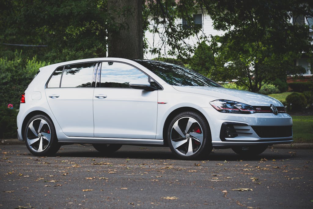 A sleek white Volkswagen GTI parked under a tree on an asphalt road, outdoors.